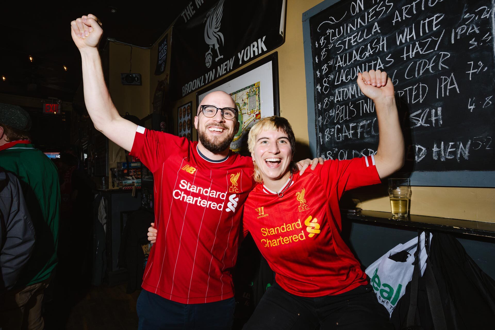 Two people raising their arms while wearing soccer shirts in a local bar